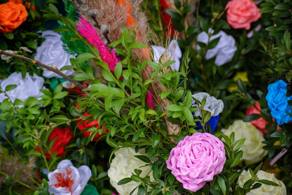 A close-up view of a vibrant floral display featuring a variety of fresh flowers including pink, white, red, blue, and orange blooms arranged together with lush green foliage. The arrangement showcase