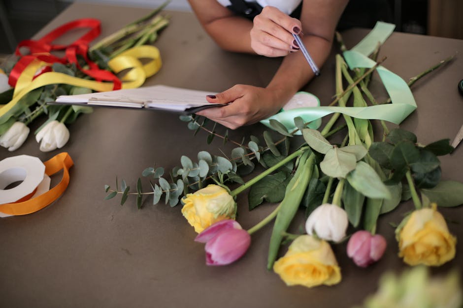 A person is working at a floral arrangement table with various fresh flowers and materials, including yellow and pink tulips, white roses, green leaves, and eucalyptus, all laid out on a brown surface