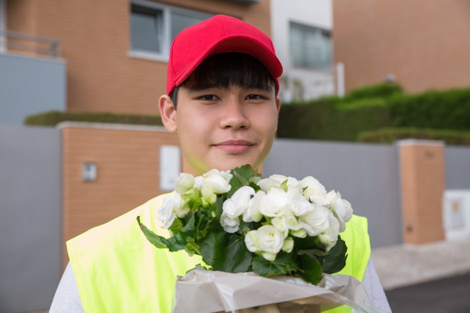 A young male flower delivery courier standing outdoors in a residential area with modern brick and concrete houses in the background. He is wearing a bright yellow high-visibility vest and a red cap, 
