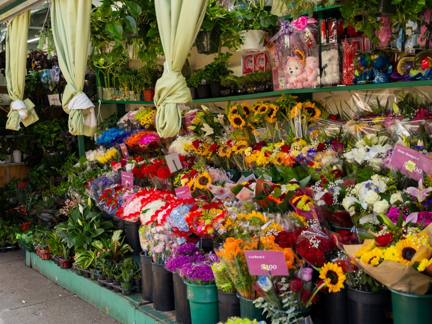 A vibrant floral display outside a flower shop featuring an array of fresh bouquets and potted plants. Prominent in the arrangement are bright yellow sunflowers, white roses, purple and pink mixed flo