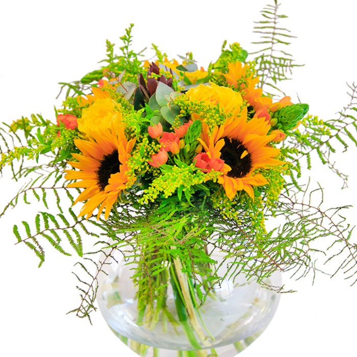 As sunlight filters softly through Hornsey's leafy avenues, this exuberant bouquet beautifully echoes the local landscape in full bloom. Anchored by radiant sunflowers with their bold, dark centers and glowing yellow petals, the arrangement beams joyfully from its perch in a perfectly round glass vase-its smooth, water-filled surface reflecting every vivid detail. Tucked between the cheerful sunflowers, luxurious yellow garden roses unfurl in tightly packed spirals, their blooms exuding a light, fresh scent that conjures late summer mornings in the Rose Garden at Priory Park. Accents of glossy red hypericum berries shimmer amid cascades of fine, airy ferns and sprays of light green goldenrod speckled with tiny yellow flowers. Eucalyptus leaves threaded throughout add a gentle, cool contrast, mirroring the subtle shifts of colour found along Green Lanes. This bouquet's texture is rich and varied: the suede-like softness of the sunflower petals, the silky fullness of the roses, the crisp greenery, and the firm, bright berries together create visual harmony and tactile pleasure. Presented by Flowers Hornsey, this arrangement is as uplifting as a walk along the New River Path-an ideal gift for birthdays, anniversaries, or any occasion requiring a dash of Hornsey happiness. Its intricate structure and glowing palette transform every setting into a place of celebration.