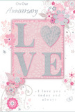 The photograph captures an anniversary card laid flat, its design glowing softly in a palette of pastel pinks, pearly whites and cool silvers that conveys a sense of long-lasting romance. At the heart of the card, the word LOVE appears in raised, glitter-soft letters, each one slightly textured and luminous, as if dusted with frost. The O is subtly suggested as a heart shape, or framed by surrounding flourishes, adding a gentle, affectionate focus to the composition. Around the central word, delicate blooms open in illustrated form, their petals shaded with blush, pale mauve and ivory, while fine silver highlights trace along some edges, similar to the refined details you might see in a bespoke anniversary bouquet created in a Hornsey studio. Slender vines and curled stems weave between the flowers, drawn with thin, graceful lines that echo the natural bend of fresh foliage arranged in a loose, romantic style. The background is a soft pink field, smooth and gently glowing, with slightly deeper tones near the centre that fade toward the borders, giving the impression of depth without any harsh contrast. Tiny accents-suggestions of petals, swirling filigree and possibly faint butterflies-add a quiet sense of movement, like blossoms stirred by a light breeze over Alexandra Park. An understated script, reading phrases such as On Our Anniversary and I love you today and always, is printed in elegant, flowing lettering that supports the design rather than dominating it. The card feels tender, thoughtful and timeless, perfect alongside a hand-delivered anniversary bouquet arriving at a doorstep anywhere around Hornsey.